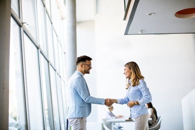 Two people shaking hands after a successful negotiation in a brightly lit office setting, with smiles on their faces.