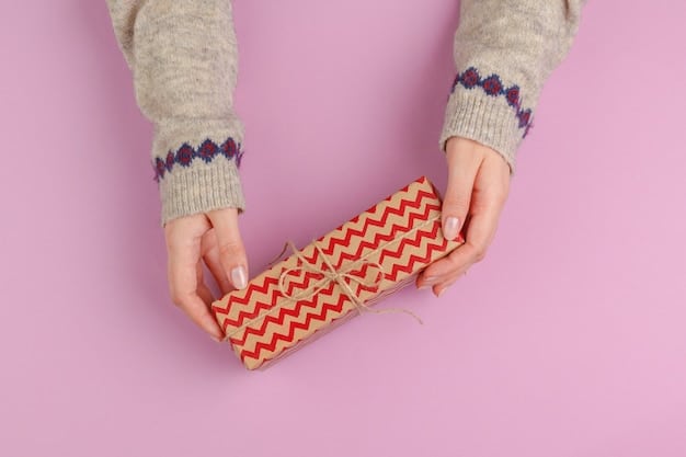 A hand carefully wrapping a small gift box with a piece of colorful patterned fabric. The fabric is being secured with twine and a small tag. The background is a wooden table with various crafting supplies scattered around, suggesting a DIY wrapping station.