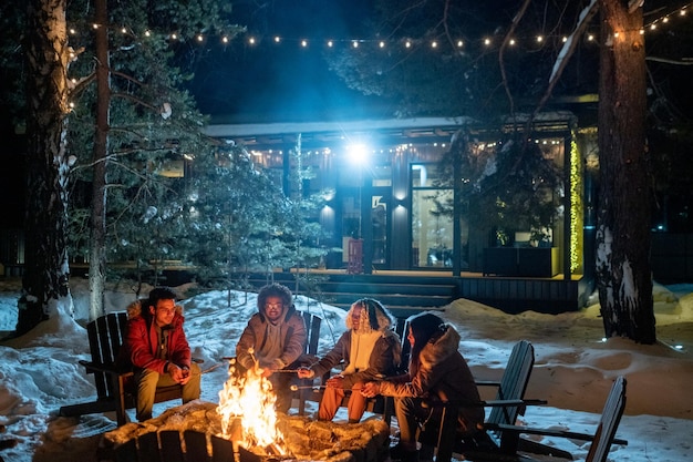An outdoor holiday party scene with friends gathered around a fire pit at dusk, surrounded by string lights and enjoying snacks and drinks.